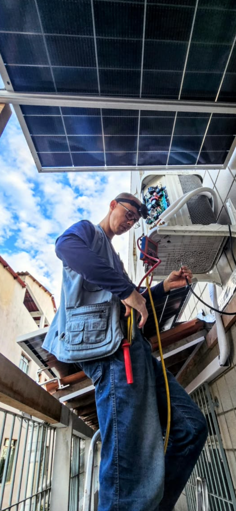 Técnico em escada realizando manutenção em condensadora de ar-condicionado sob cobertura de painéis solares
